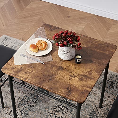 Wooden table with a plate of buns and a vase of red flowers.